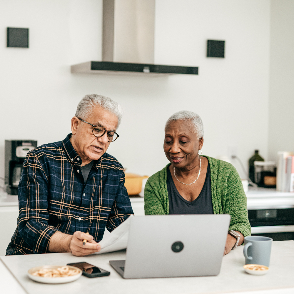 An older couple sitting at a kitchen table, reviewing documents on a laptop together.