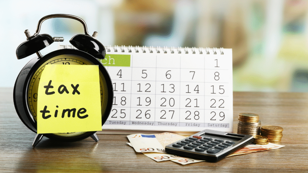 Black alarm clock with a sticky note saying 'tax time' next to a calendar and coins.